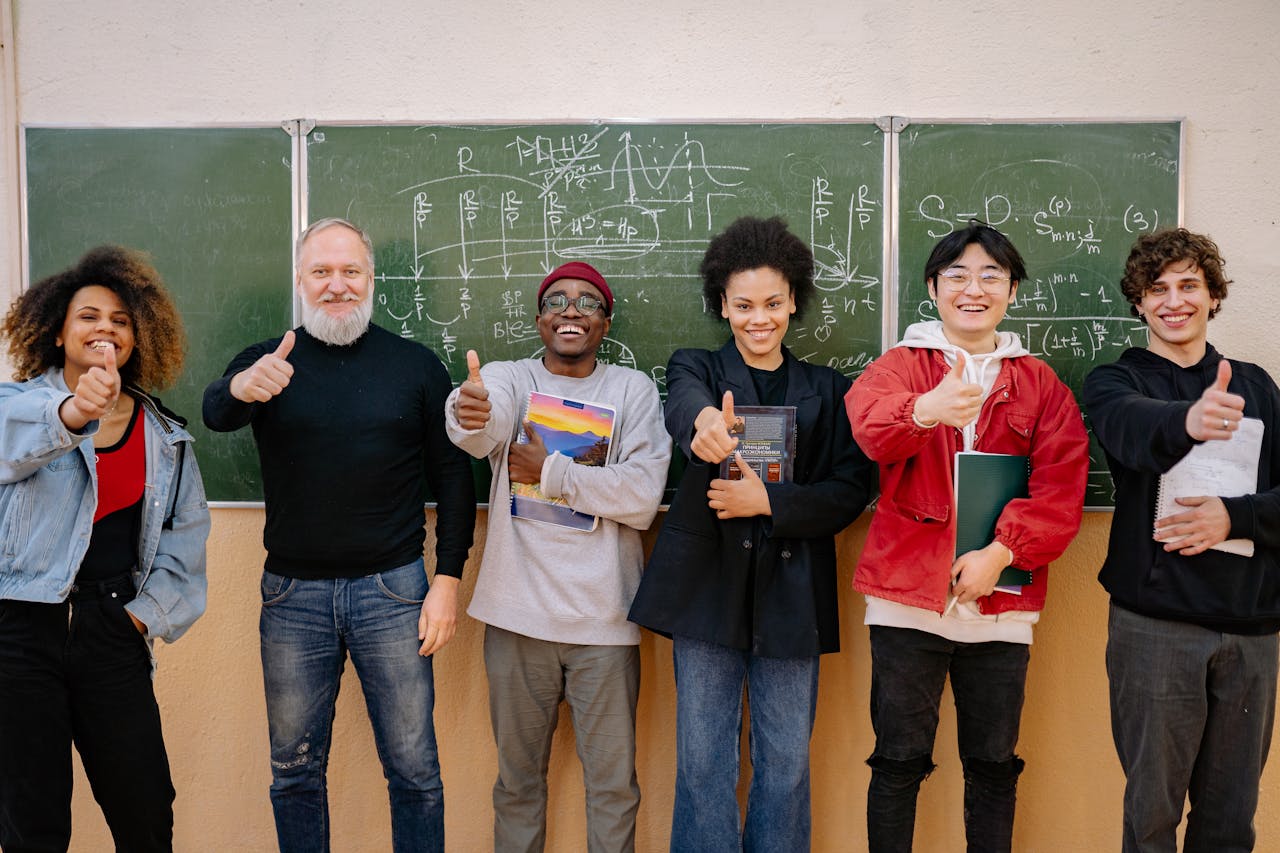 gallery-2 A group of diverse students with a teacher, all thumbs up, in front of a chalkboard with equations.